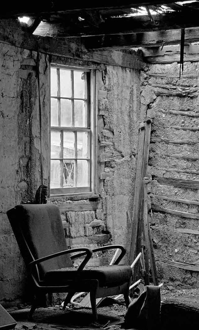 A worn armchair beneath the window of a ruined stone cottage, in black and white.